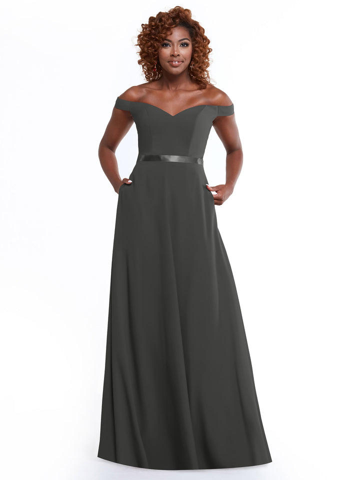 A woman with curly hair smiles in the Avery Austin 80100 dress—an off-the-shoulder, floor-length, dark gray A-line gown with a fitted bodice, satin waistband, and pockets—standing against a plain white background.