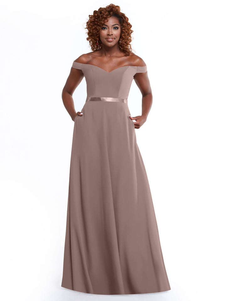 A woman with curly hair models Avery Austin’s 80100 off-the-shoulder, floor-length A-line dress in mauve with a satin waistband and pockets, standing before a plain white backdrop.