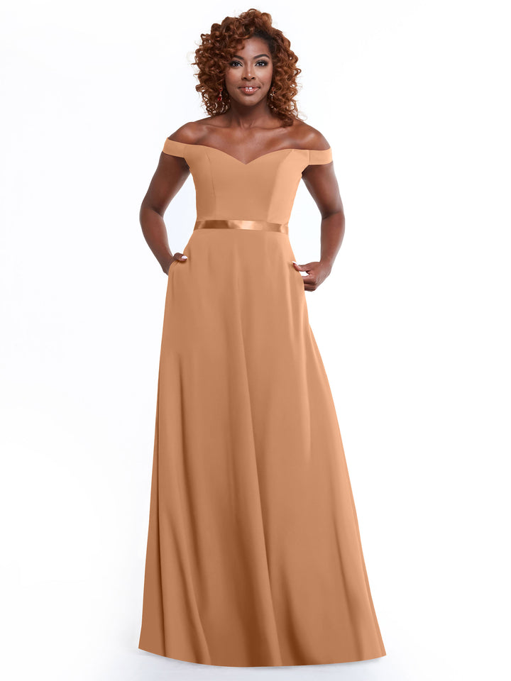 A woman with curly hair models the Avery Austin 80100, a floor-length light brown satin off-the-shoulder A-line dress with a waistband and pockets, standing against a plain white background.