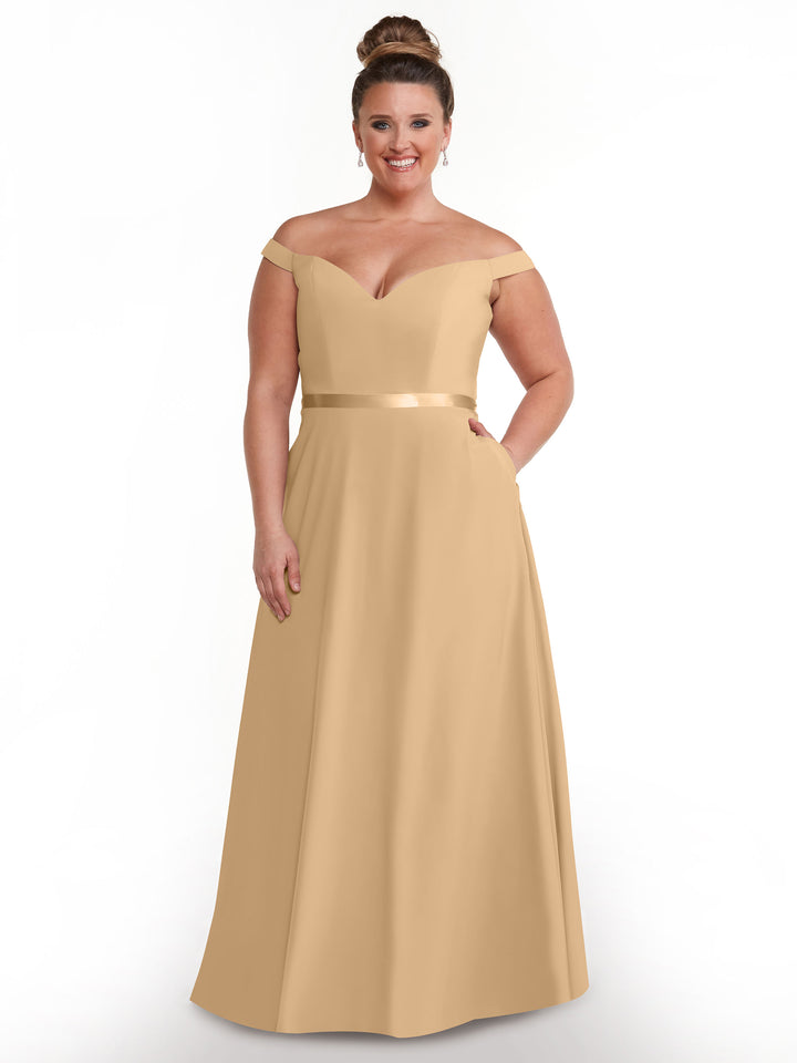 A woman stands smiling against a white background in Avery Austin’s 80100—a long, off-the-shoulder A-line dress with a satin waistband and pockets. Her hair is in an elegant bun and she wears drop earrings.