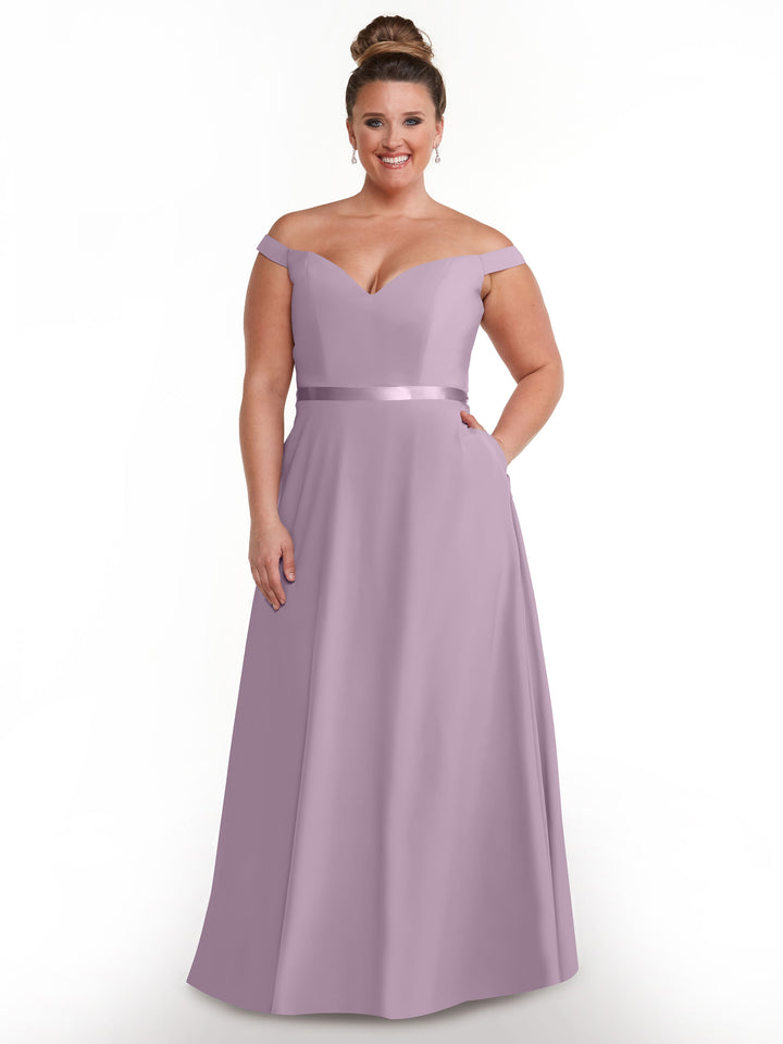 A woman in the Avery Austin TH-80100, a dusty lavender floor-length A-line dress with an off-the-shoulder neckline and satin waistband, stands smiling against a plain white background. Her hair is in a neat bun, accented by drop earrings.