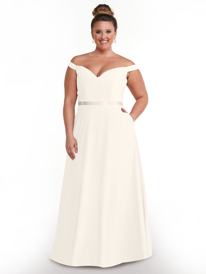 A woman stands against a white background, smiling in Avery Austin’s style 80100: an off-the-shoulder, floor-length A-line dress with a V-neckline and satin waistband. Her hair is elegantly styled in a bun.