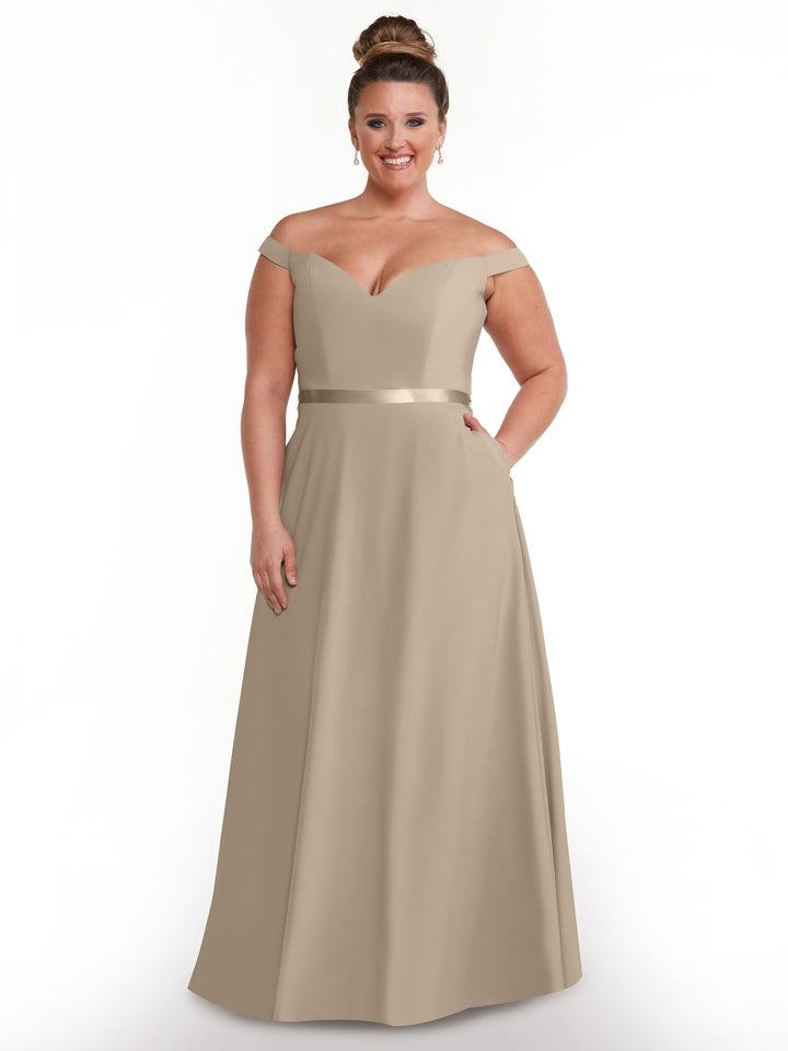 A woman wearing the Avery Austin 80100, an elegant off-the-shoulder A-line dress in taupe with a satin waistband, stands and smiles against a plain white background.