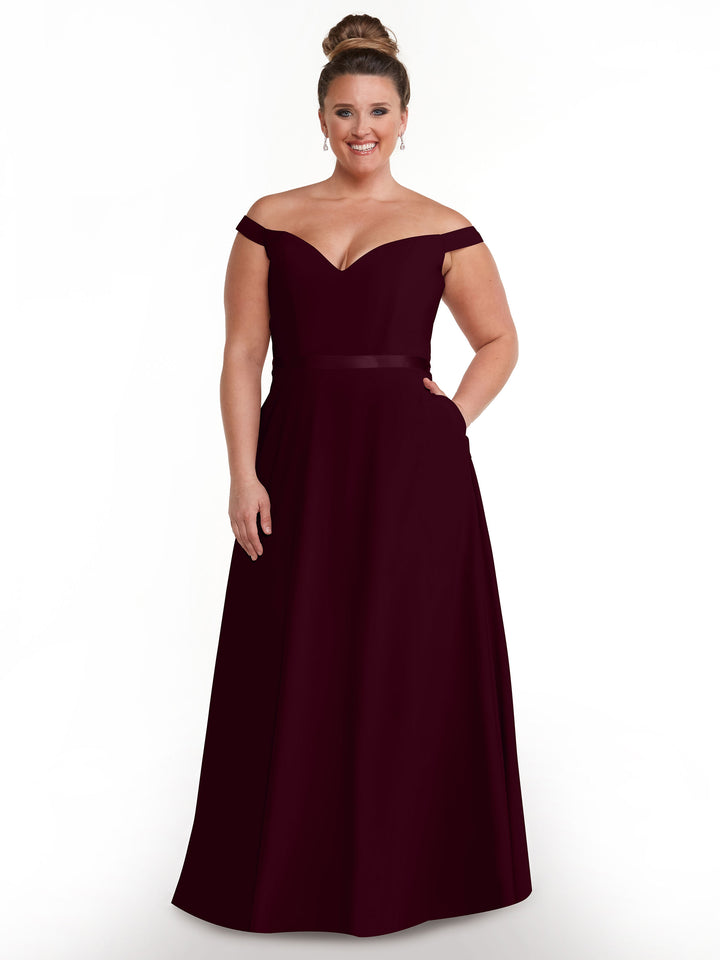 A woman wearing the Avery Austin TH-80100 maroon, off-the-shoulder dress stands smiling against a white background. Her floor-length gown is styled with a high bun and elegant drop earrings.