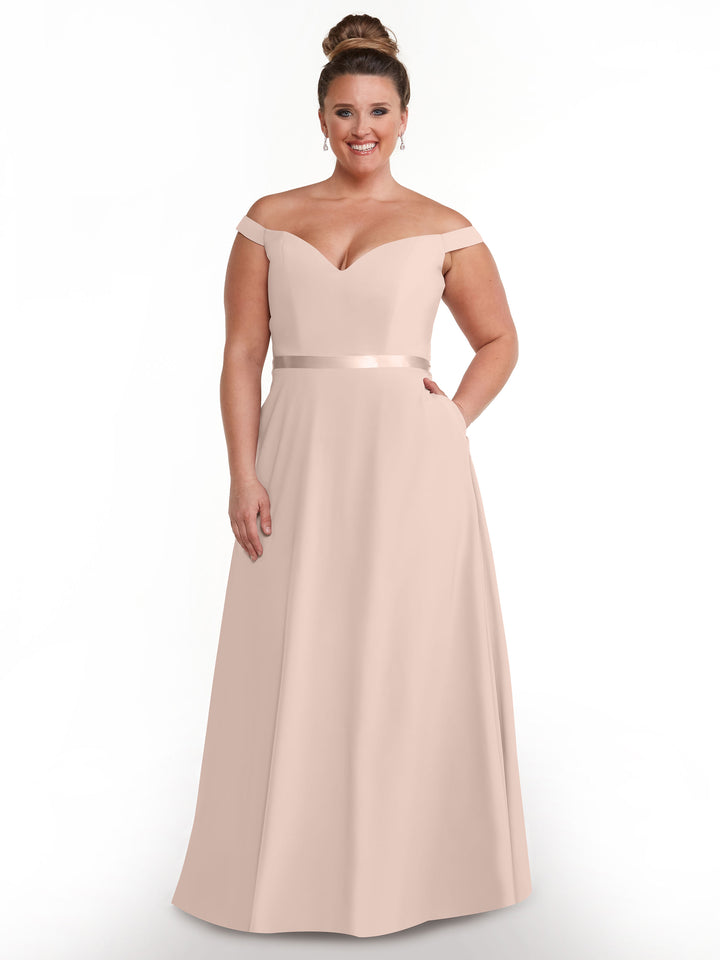 A woman models the Avery Austin 80100, a blush pink off-the-shoulder gown with a sweetheart neckline and satin waistband, standing against a white background to highlight its elegant, floor-length style.