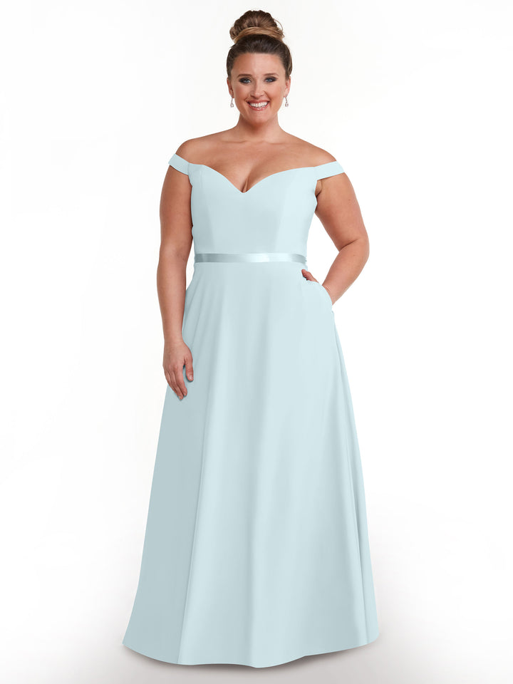 A woman smiles in the Avery Austin 80100, a timeless light blue off-the-shoulder dress with a satin waistband and side pockets, her hair in a neat bun as she poses against a plain white background.