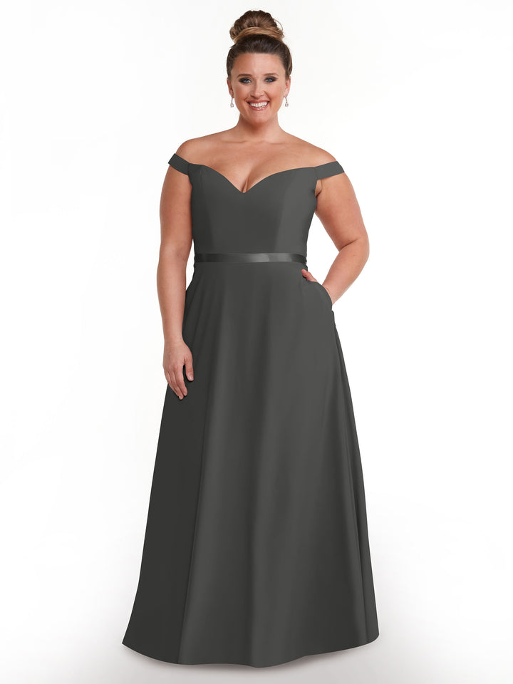 A smiling woman with light brown hair in a bun wears the Avery Austin 80100, a timeless dark gray off-the-shoulder, floor-length A-line dress, standing against a plain white background.