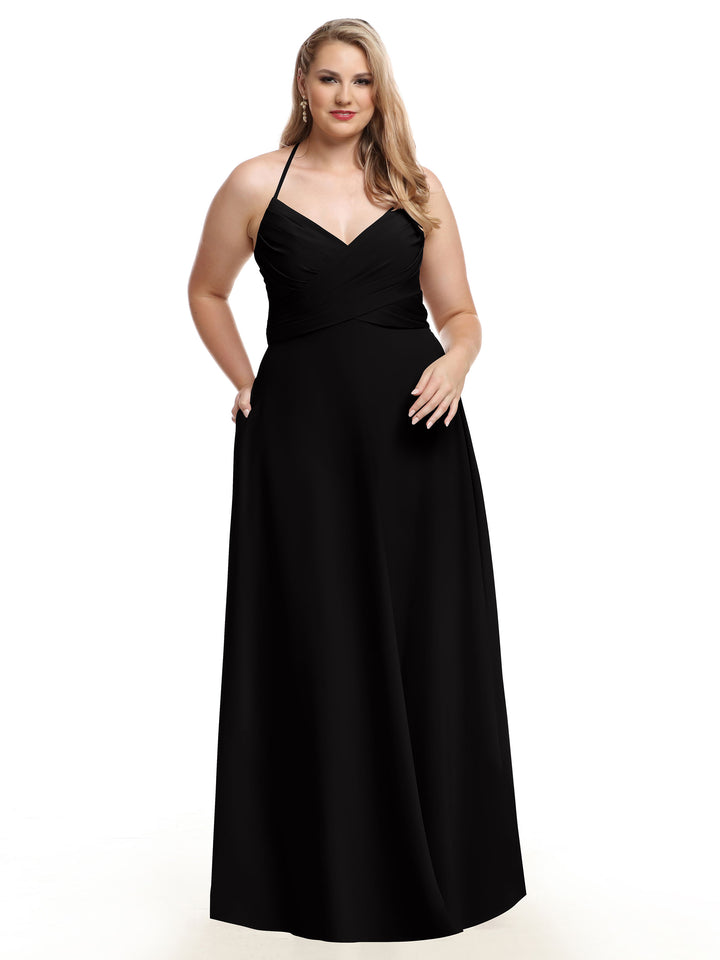 A woman models Avery Austin’s 80101, a long black A-line dress with a pleated bodice and V-neckline, standing against a white background and smiling softly with one hand in her pocket.