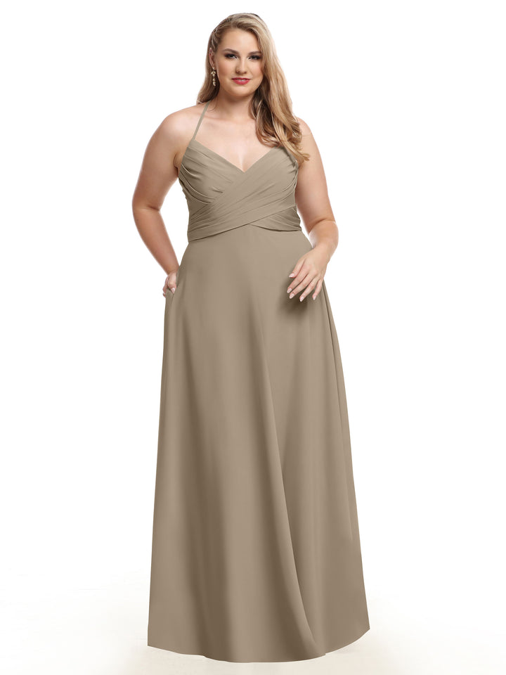 A woman wears the Avery Austin 80101, a taupe floor-length A-line dress with a fitted pleated bodice and spaghetti straps, posing with one hand in her pocket against a white background.