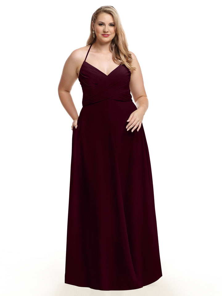 A woman with long, wavy blonde hair stands against a white background wearing Avery Austin’s 80101: a sleeveless, floor-length dark burgundy A-line dress with a halter neckline, one hand in her pocket and the other by her side.