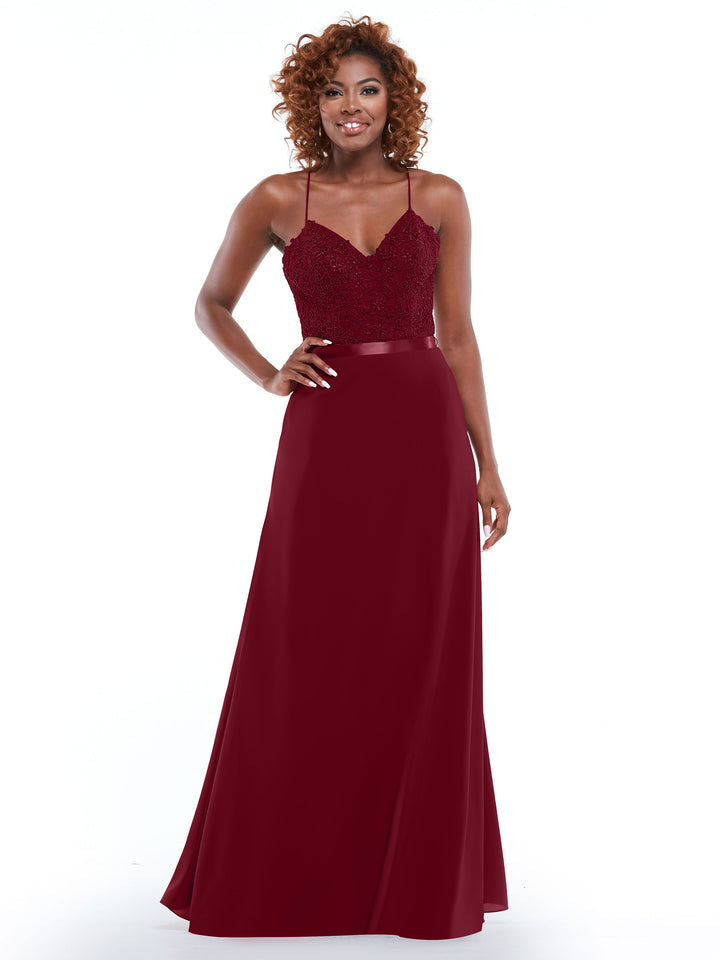 A woman with curly hair is smiling in the Avery Austin TH-80103—a floor-length, burgundy spaghetti strap gown with a fitted lace bodice and satin waistband—while standing against a plain white background.