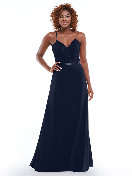 A woman with curly hair smiles against a white background, wearing the Avery Austin 80103—a dark navy evening gown with spaghetti straps, a lace bodice, and a long, flowing A-line skirt with a slit.