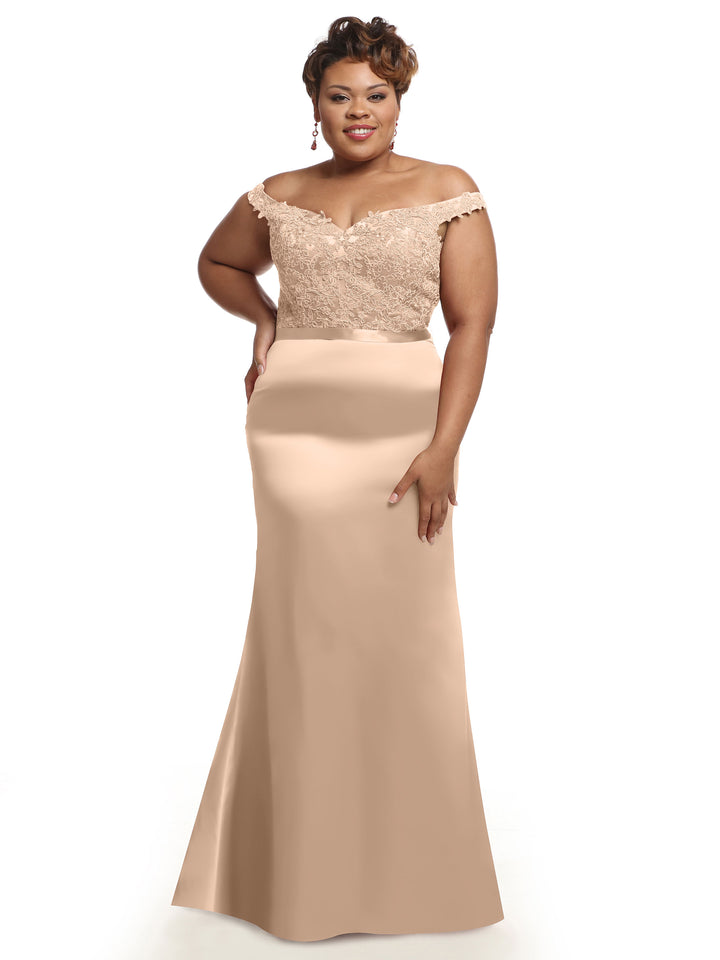 A woman smiles warmly against a white background, wearing Avery Austin’s 80104 bridesmaid dress with a lace bodice and smooth satin, floor-length skirt in champagne. She has short hair and accessorizes with earrings.