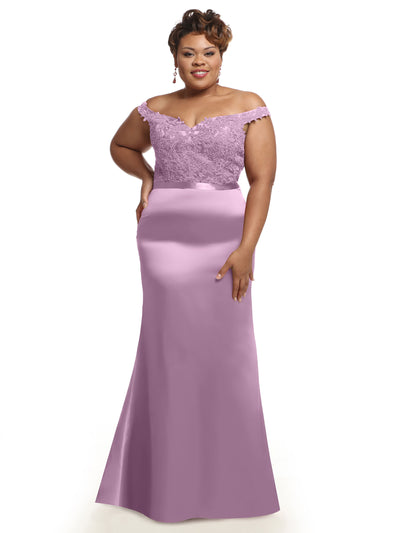 A woman stands smiling with one hand on her hip, wearing the Avery Austin 80104 dress in lilac, featuring an off-the-shoulder lace bodice and a flowing satin skirt, against a white background.