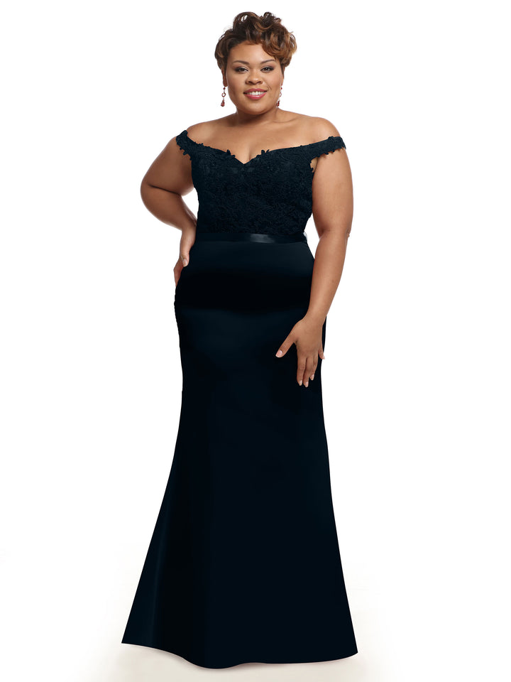 A woman poses confidently in Avery Austin’s 80104, a long off-the-shoulder bridesmaid dress featuring a lace bodice, with one hand on her hip against a plain white background.