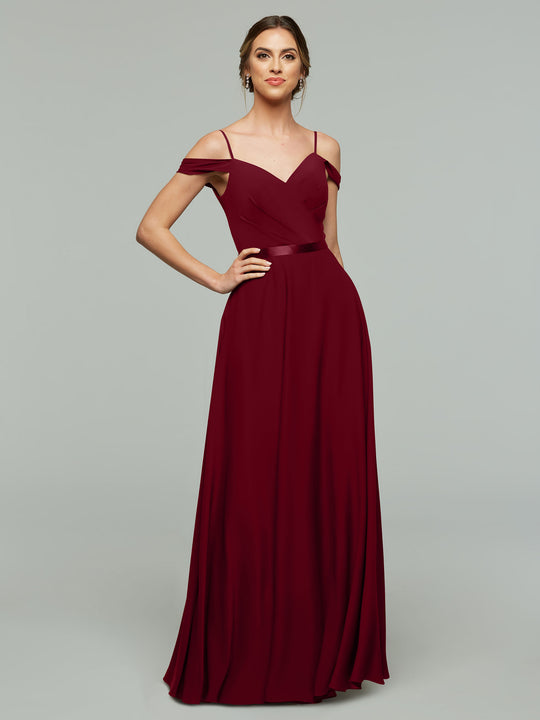A woman models Avery Austin’s 80106: a burgundy chiffon, floor-length bridesmaid dress with thin straps, an off-the-shoulder fitted bodice, and flowing skirt, posing against a plain gray background with one hand on her hip.