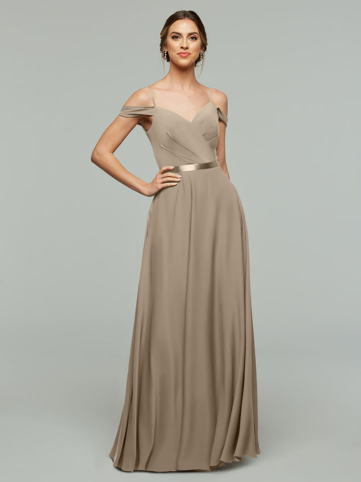 A woman models Avery Austin style 80106, a long taupe off-the-shoulder bridesmaid dress with draped sleeves, ruched bodice, and flowing skirt, standing with one hand on her hip against a light gray background.