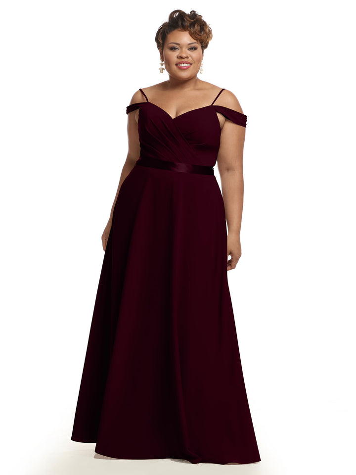 A woman smiles in the Avery Austin 80106 maroon floor-length, off-the-shoulder bridesmaid dress with a fitted bodice and flowing skirt. She has short hair, wears dangling earrings, and stands against a white background.