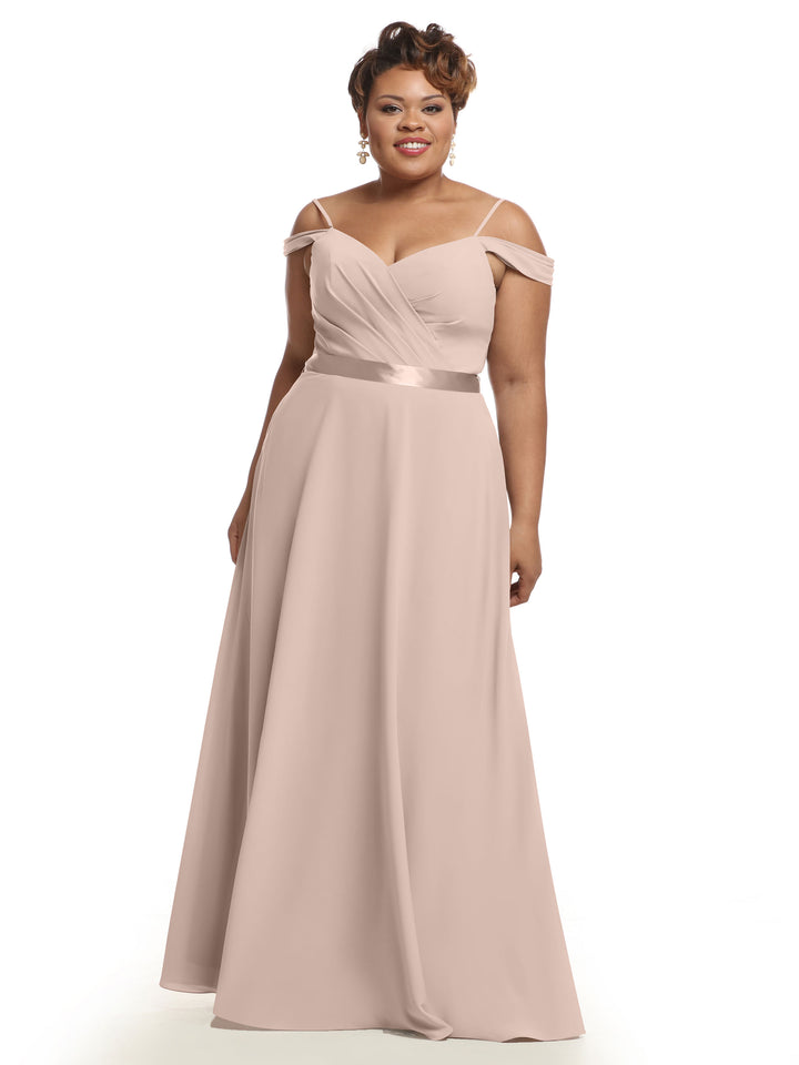 A woman smiles in the Avery Austin 80106 bridesmaid dress, featuring a rose pink hue, off-the-shoulder V-neckline, fitted bodice, and flowing skirt. She has short hair, dangling earrings, and stands against a white background.