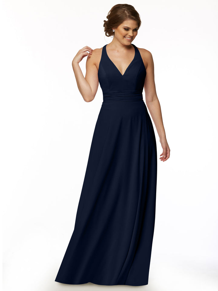 Wearing Avery Austin’s 80107, a sleeveless, floor-length navy A-Line dress with a deep V-neck, a woman stands against a white background, smiling and touching her hair, radiating graceful feminine charm.