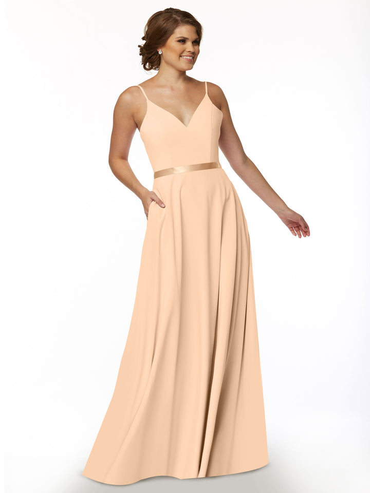 A woman models Avery Austin’s 80112, a long peach A-line dress with a V-neck, spaghetti straps, and a satin waist sash, standing against a plain white background.