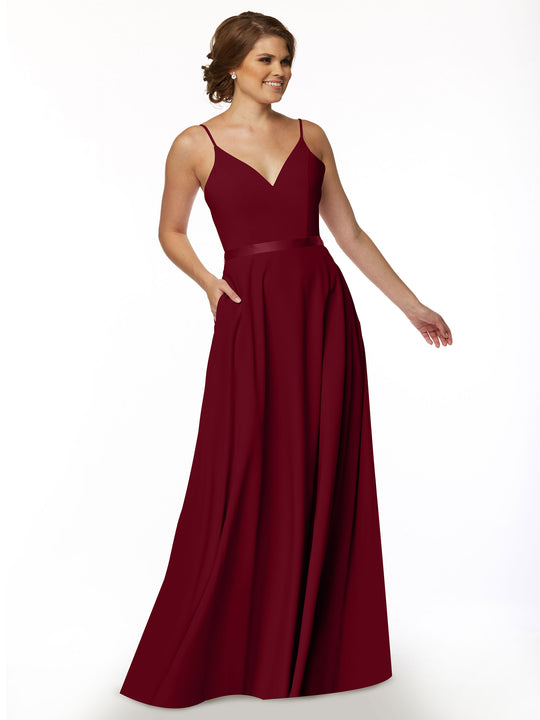 A woman models Avery Austin’s 80112: a sleeveless, floor-length burgundy A-line dress with a V-neck and fitted waist, posing with one hand in her pocket and smiling against a plain white background.