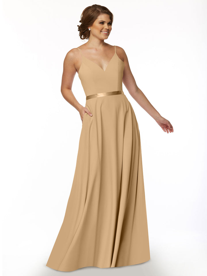 A woman wearing Avery Austin style 80112, a tan A-line dress with V-neck, satin sash waistband, and pockets, stands smiling against a white background.