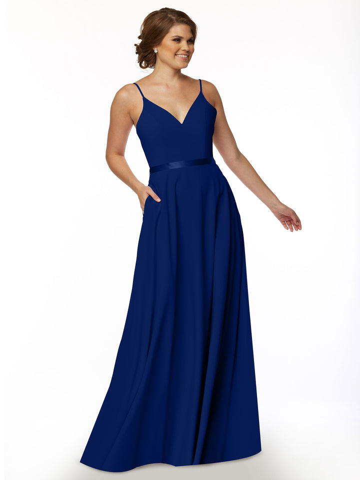 A woman wears Avery Austin’s 80112, a sleeveless floor-length deep blue A-line dress with a V-neckline and satin waistband, standing against a plain white background.