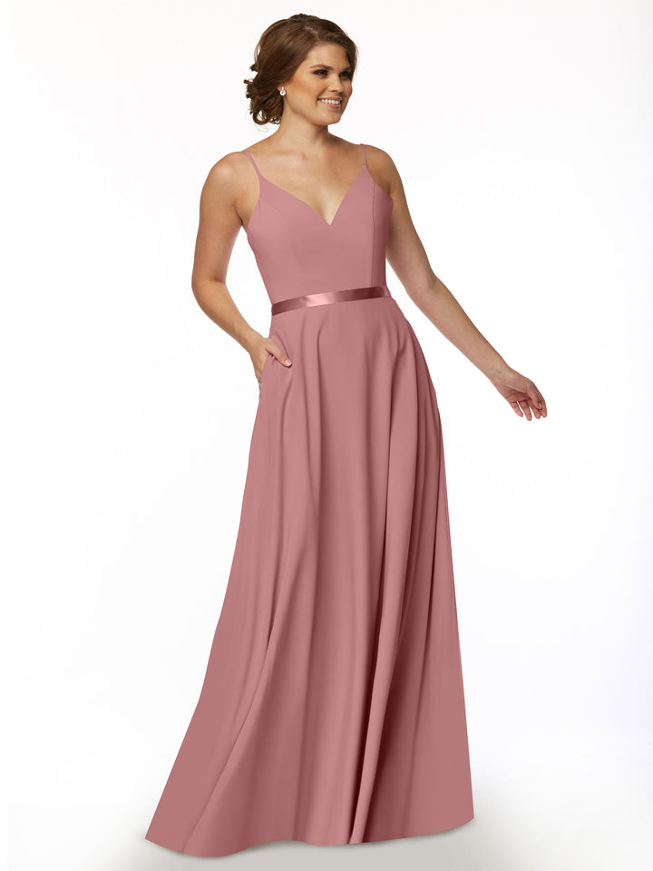 A woman smiles with one hand in her pocket, wearing the Avery Austin 80112— a sleeveless, floor-length dusty rose A-line dress with a V-neckline and satin sash waistband— against a plain white background.