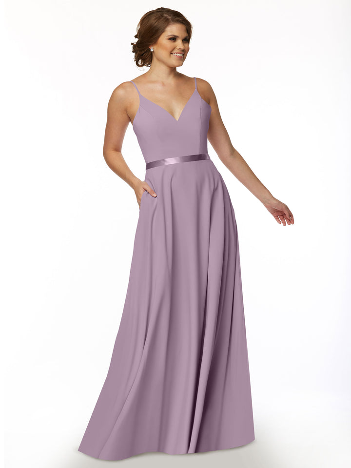 A woman wearing the Avery Austin TH-80112, a long sleeveless lavender A-Line dress with a V-neckline and satin sash, stands against a white background, smiling with her hands in the dress pockets.