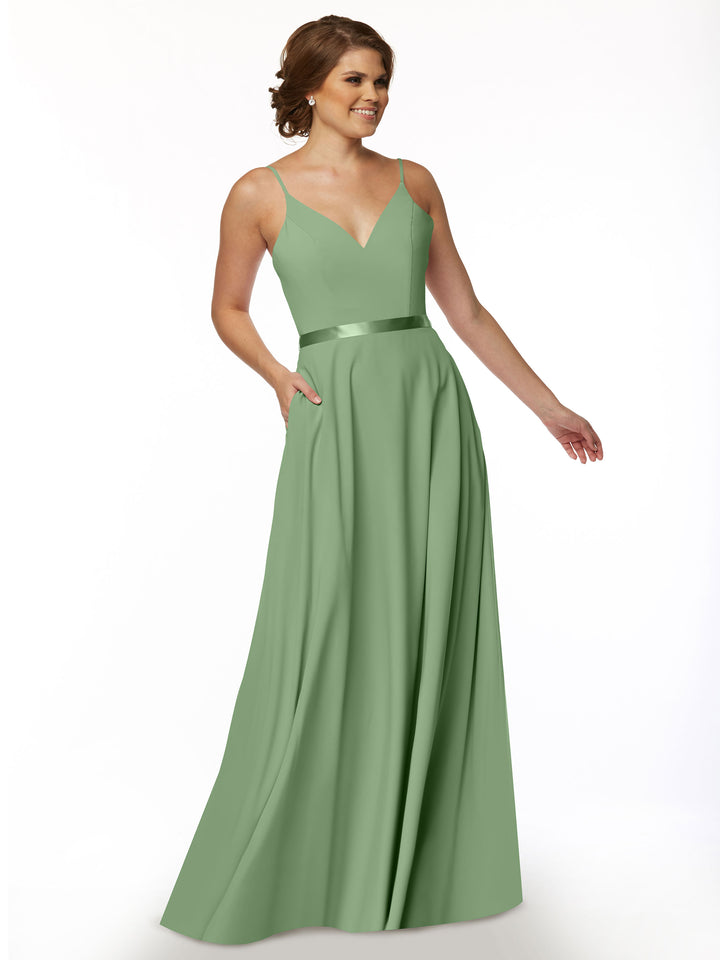 A woman wearing Avery Austin’s 80112—a long sage green A-line dress with spaghetti straps, V-neckline, satin sash waistband, and pockets—stands against a white background, smiling with one hand in her pocket.