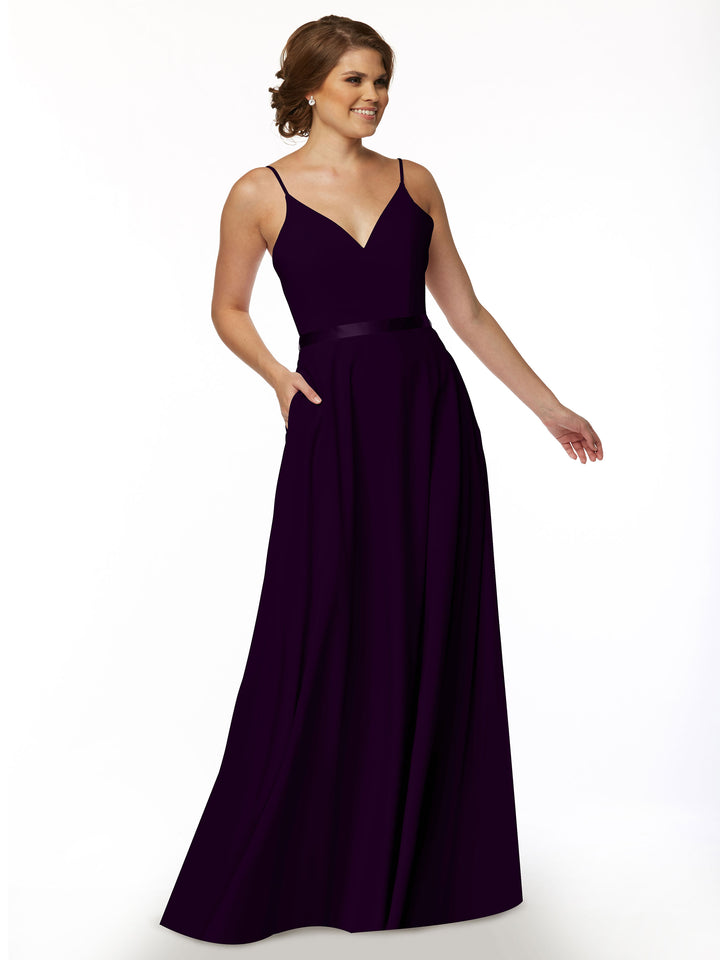 A woman stands smiling with one hand in her pocket, wearing Avery Austin’s 80112—a sleeveless deep purple A-line dress featuring a V-neckline and spaghetti straps—against a plain white background.