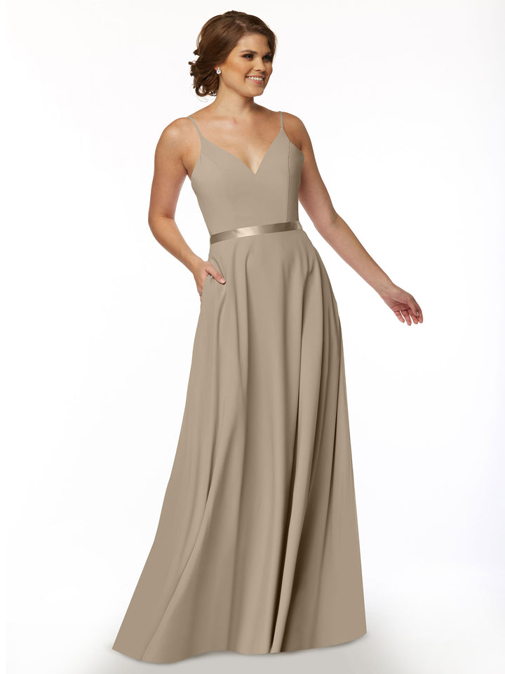 A woman stands smiling with one hand in her pocket, wearing Avery Austin’s 80112—a long taupe A-line dress with spaghetti straps, a V-neckline, satin sash waistband, and flowing skirt that complements her styled hair and earrings.