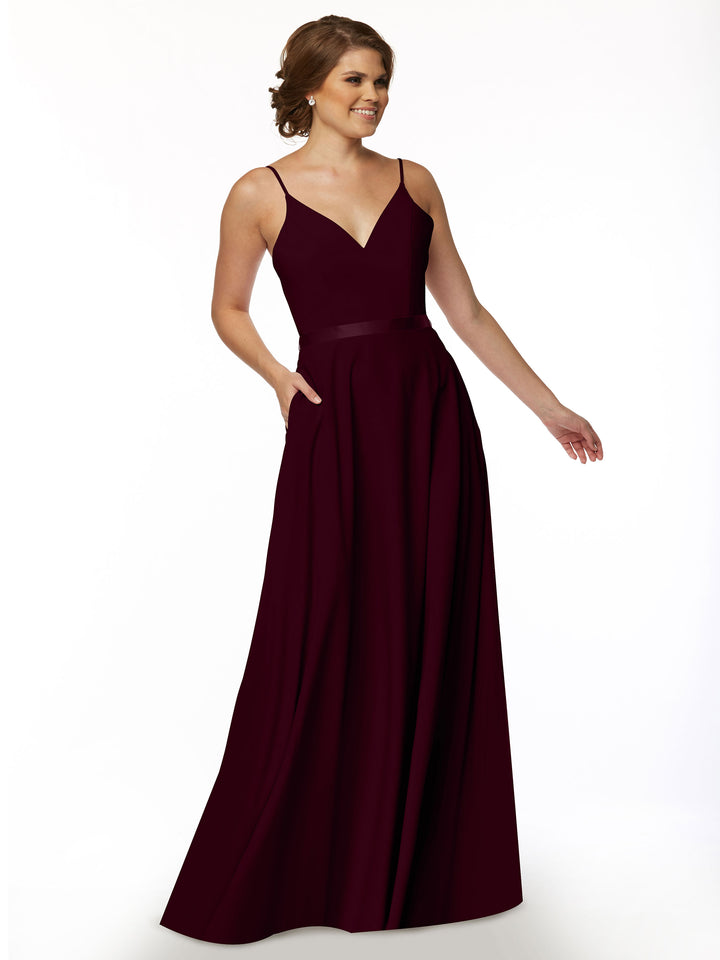 A woman models Avery Austin’s 80112, a sleeveless deep burgundy floor-length A-line dress with spaghetti straps, V-neckline, and pockets, standing against a plain white background.