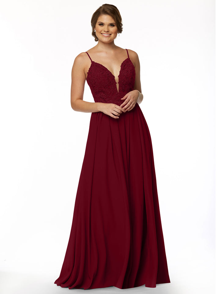 A woman in Avery Austin style 80113, a sleeveless floor-length burgundy gown featuring an illusion plunge neckline, lace bodice, and flowing A-line chiffon skirt, stands smiling against a plain white background.