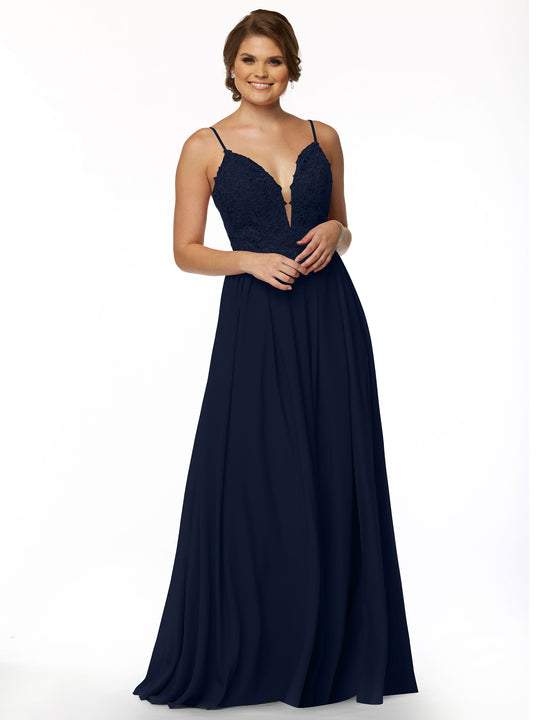 A woman stands smiling against a plain white background, wearing Avery Austin’s 80113: a sleeveless navy blue evening gown with an illusion plunge neckline, lace bodice, and thin straps.