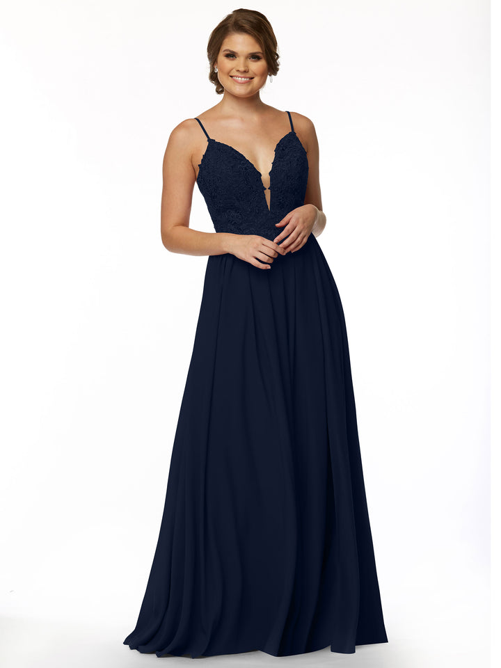 A woman stands smiling against a plain white background, wearing Avery Austin’s 80113: a sleeveless navy blue evening gown with an illusion plunge neckline, lace bodice, and thin straps.