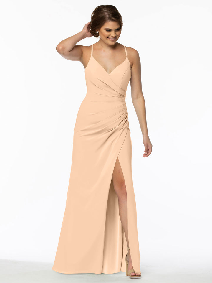 A woman models Avery Austin style 80114, a sleeveless light peach chiffon bridesmaid dress with a v-neckline, draped waist, and thigh-high slit, smiling in heels with one hand touching her hair.