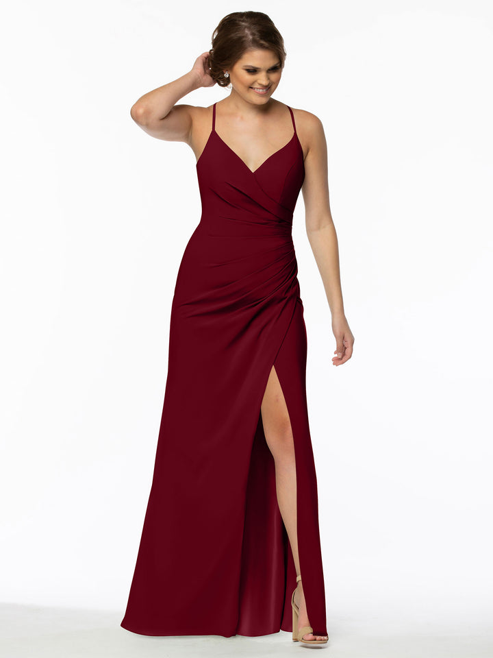 A woman wearing Avery Austin’s 80114 sleeveless, floor-length burgundy chiffon bridesmaid dress with a thigh-high slit stands against a white background, smiling and touching her hair while wearing nude high-heeled sandals.