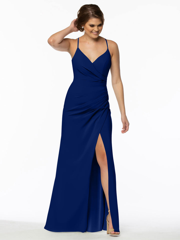 A woman wears Avery Austin style 80114, a sleeveless deep blue v-neck bridesmaid dress with thin straps, fitted bodice, and high leg slit. She smiles with one hand in her hair against a plain white background.