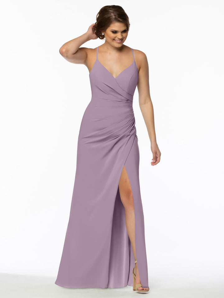 A woman in the Avery Austin 80114, a sleeveless mauve chiffon bridesmaid dress featuring a V-neckline, ruched waist, and high slit, stands against a white background smiling with one hand in her hair.