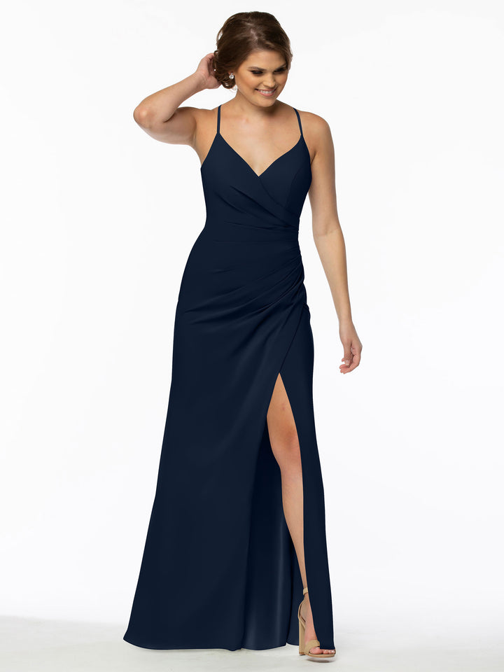 A woman smiles and touches her hair while wearing the Avery Austin 80114 navy blue v-neck bridesmaid dress, featuring thin straps, a wrap-style bodice, and a thigh-high slit, standing against a white background.