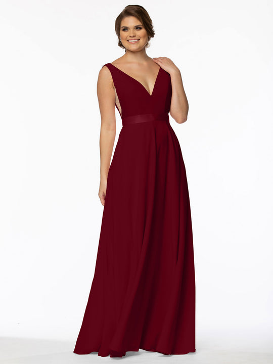 A woman models Avery Austin’s 80117: a sleeveless, deep V-neck, floor-length burgundy gown with illusion sides, smiling and touching her neck against a plain white background.