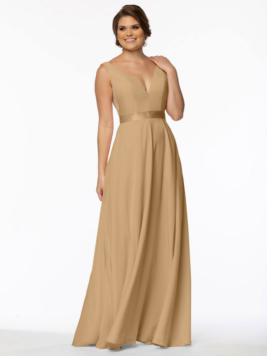 A woman with short brown hair smiles in Avery Austin’s 80117: a tan, sleeveless, floor-length evening gown featuring an A-Line silhouette, deep V-neckline, and fitted bodice. She poses with one hand raised to her neck against a plain white background.