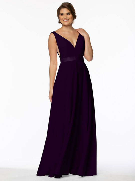 A woman smiles against a white background, touching her shoulder while wearing Avery Austin's 80117—a floor-length deep purple A-line gown featuring a plunging V-neckline and fitted waistband.