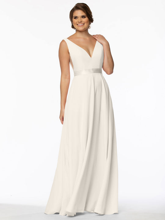 A woman wearing Avery Austin’s 80117, a sleeveless floor-length white A-Line gown with a deep V-neck and illusion sides, stands against a plain white background, smiling with one hand touching her neck.
