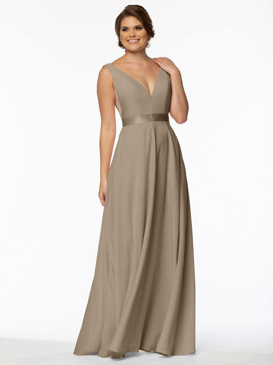A woman smiles in Avery Austin's 80117 gown—a sleeveless, floor-length taupe dress with a deep V-neckline, illusion plunging sides, a satin waistband, and an elegant A-line silhouette—posed against a plain white background.