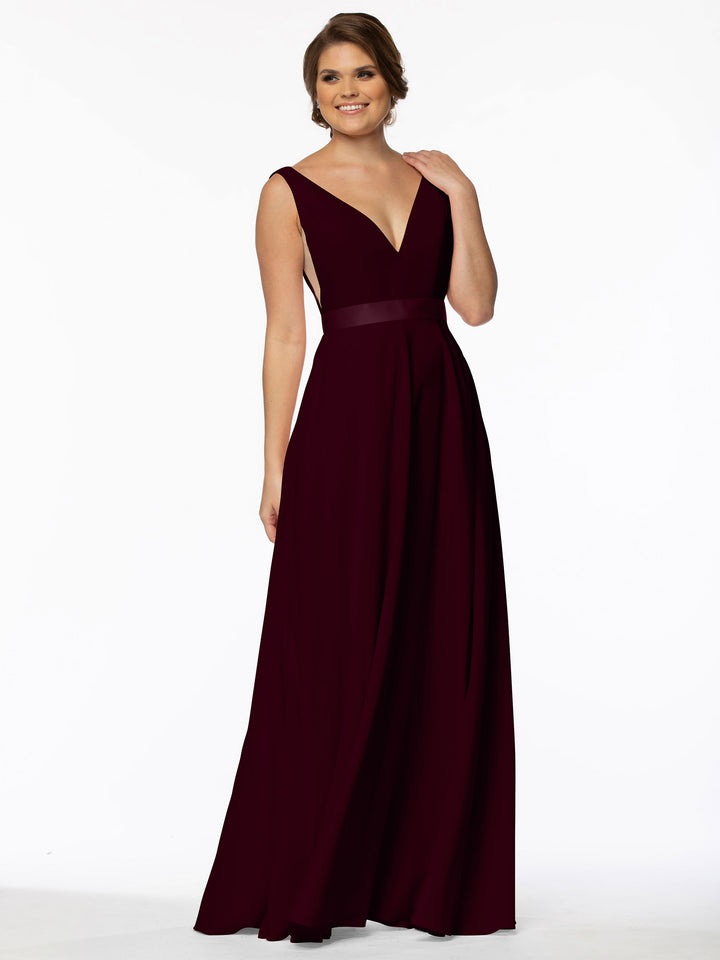 A woman poses with a soft smile and hand in her hair, wearing the Avery Austin 80117, a sleeveless A-Line gown in burgundy with floor-length hem and illusion plunging sides, set against a plain white background.