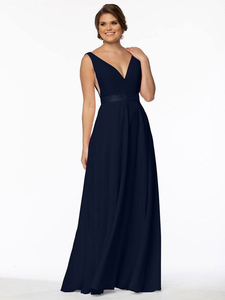 A woman smiles against a white background wearing Avery Austin gown 80117, a navy blue, floor-length A-line dress with a sleeveless design, deep V-neckline, and fitted waist.