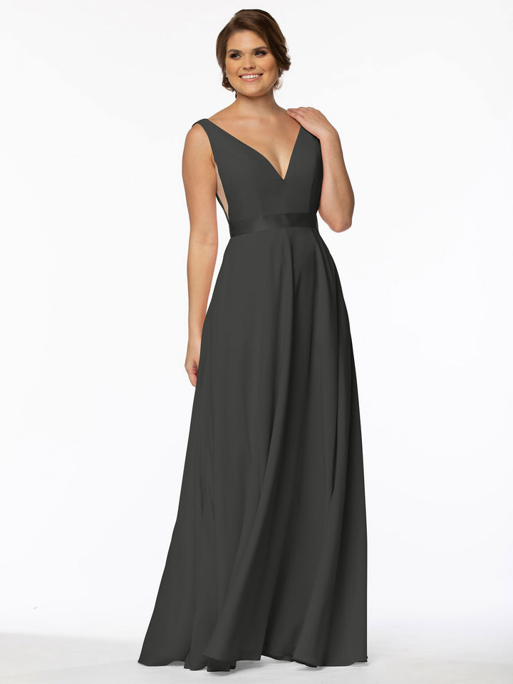 A woman smiles gently with one hand to her neck, wearing Avery Austin’s 80117—an A-line, floor-length sleeveless gown in dark gray with a deep V-neck, standing against a plain white background.